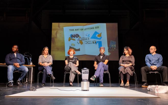 Six people sitting on a stage. In the central foreground a blender, in the background a collage.