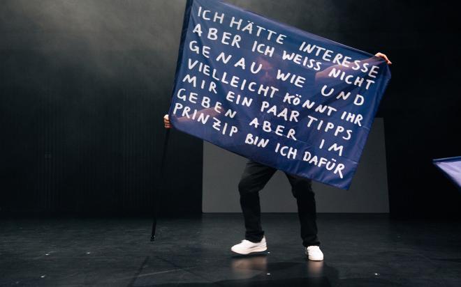 Eine Person steht auf einer dunklen Bühne und hält eine große blaue Fahne an einem schwarzen Stab. Die Fahne trägt in weißer Handschrift den Text: „Ich hätte Interesse aber ich weiss nicht genau wie und vielleicht könnt ihr mir ein paar Tipps geben aber im Prinzip bin ich dafür." Der Oberkörper der Person ist hinter der Fahne verborgen, sichtbar sind nur Beine in dunkler Hose und weiße Sneaker. Der Bühnenhintergrund ist dunkel mit leichtem Nebel.