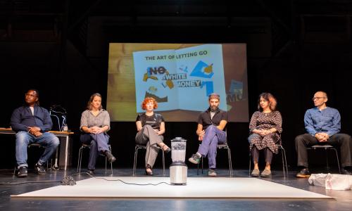 Six people sitting on a stage. In the central foreground a blender, in the background a collage.